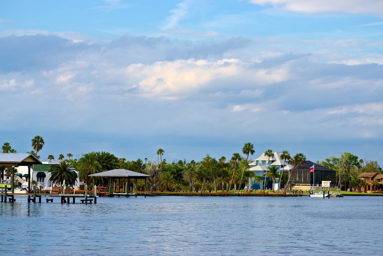 A calm waterfront scene with docks, palm trees, waterfront houses, and partly cloudy skies reflecting on the water.