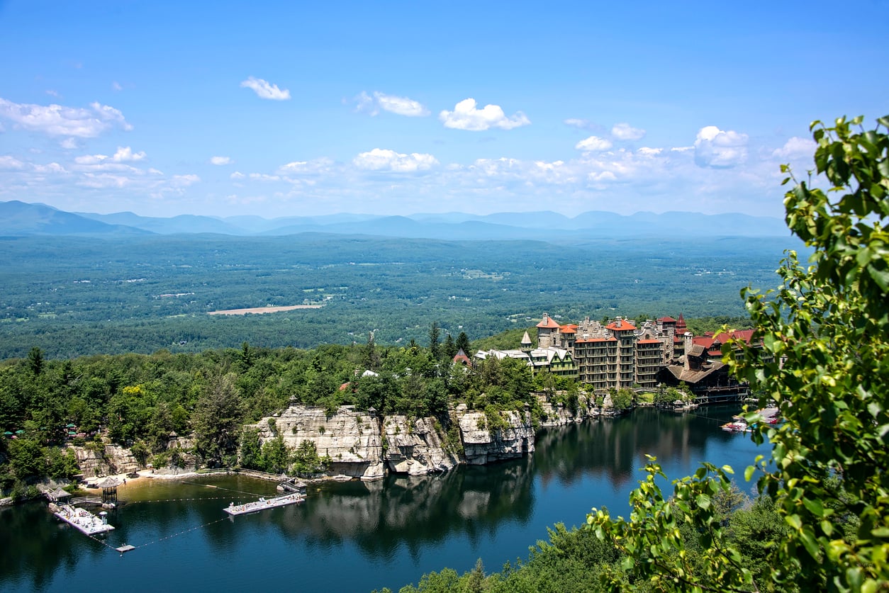 A large lakeside resort, part of the Historic Hotels collection, sits atop rocky cliffs, surrounded by dense green forest and mountains under a blue sky with scattered clouds.