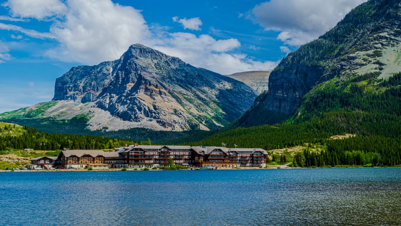 A large rustic lodge, part of the region’s renowned Historic Hotels, sits by a blue lake, surrounded by pine trees and dramatic rocky mountains under a partly cloudy sky.