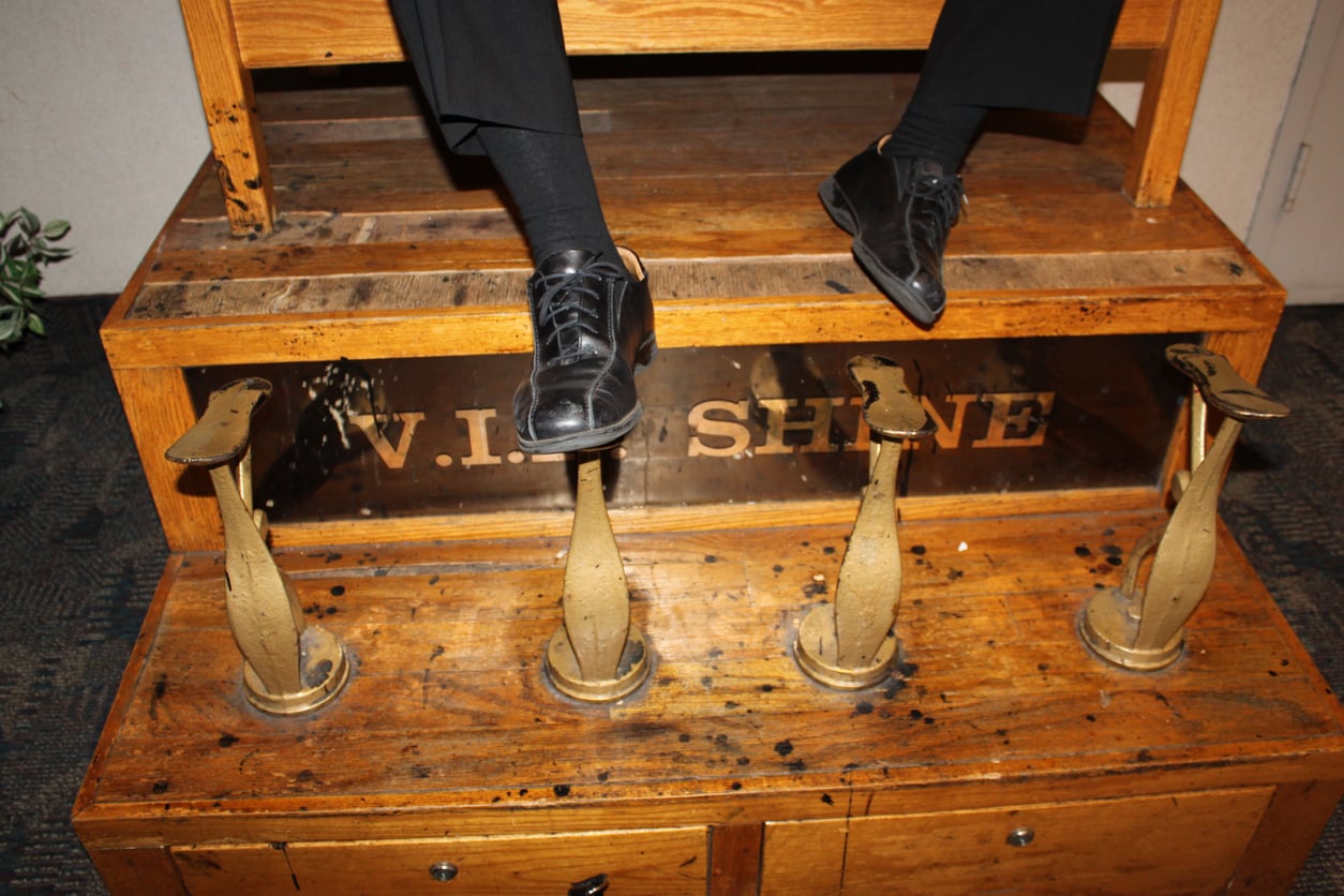 A person’s feet in black dress shoes rest on a wooden shoe shine stand labeled “V.I.P. SHINE,” with four metal footrests visible on the platform below—one of those timeless things hotels used to offer guests for a touch of classic service.