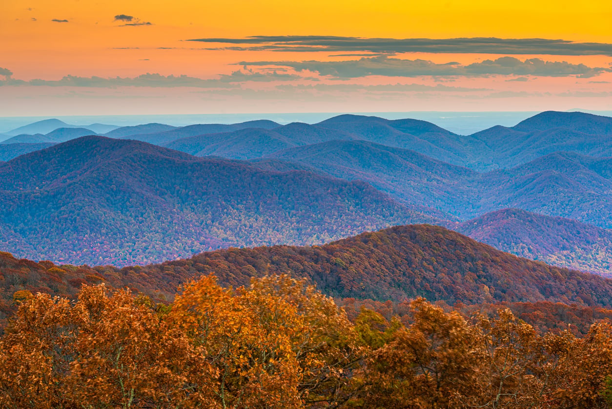 Colorful sunset over rolling blue and purple mountains with orange autumn trees in the foreground evokes the breathtaking beauty found in some of the best fall towns in America, all beneath a clear sky with scattered clouds above.