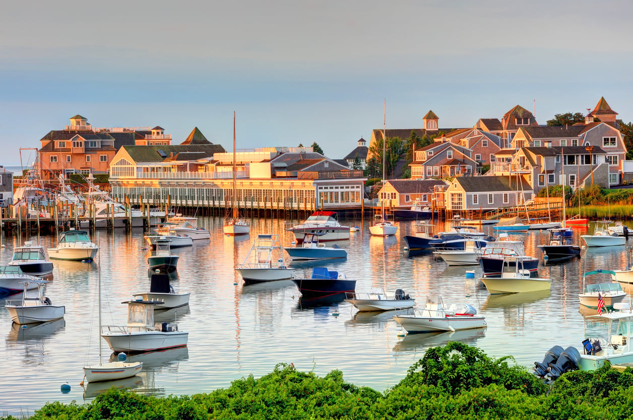 Boats are docked in a calm harbor with wooden buildings and shingled houses behind, all bathed in warm sunlight. Green shrubs are in the foreground, making this peaceful setting one of the most inviting vacation spots for relaxation and scenic beauty.