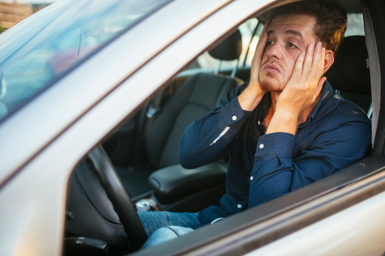 A man sits in the driver’s seat of a car with his hands on his face, looking frustrated or stressed, gazing out the window.