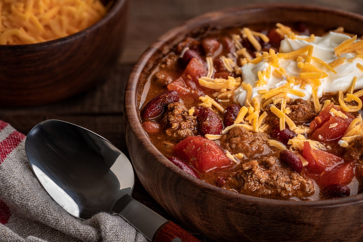 A wooden bowl of chili with ground beef, red beans, tomato chunks, shredded cheddar cheese, and a dollop of sour cream, next to a spoon and a cloth napkin.