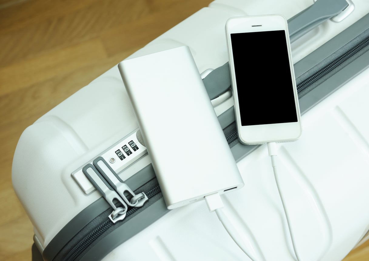 A white smartphone charging from a portable power bank placed on top of a closed white suitcase with a combination lock, on a wooden floor.
