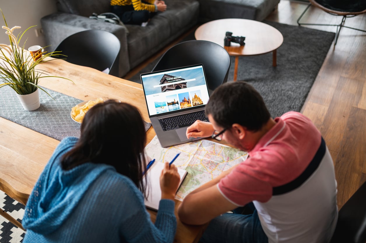 Two people sit at a wooden table, looking at a map and writing notes as they discuss their travel behaviors. A laptop with travel images is open in front of them. A bowl of chips, a plant, and living room furniture are also visible in the background.
