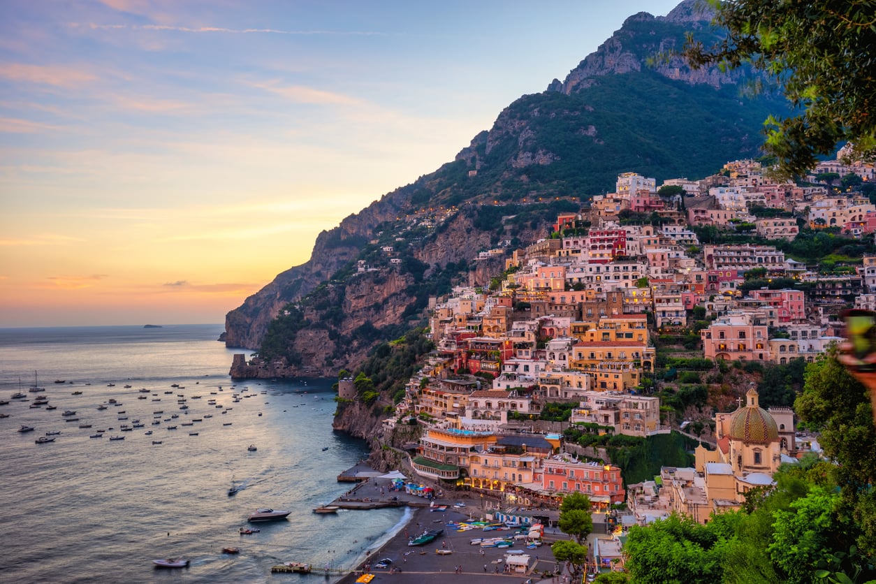 Colorful cliffside buildings overlook a bay filled with boats at sunset in Positano, Italy; mountains and calm sea frame the scenic coastal village, making it a picture-perfect spot—despite being one of the Travel Destinations Boomers Are Skipping.