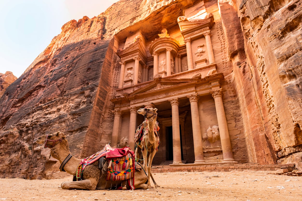 Two camels with colorful saddles rest in front of the ancient rock-carved Al-Khazneh (The Treasury) in Petra, Jordan, under warm sunlight. The intricate sandstone facade rises behind them.