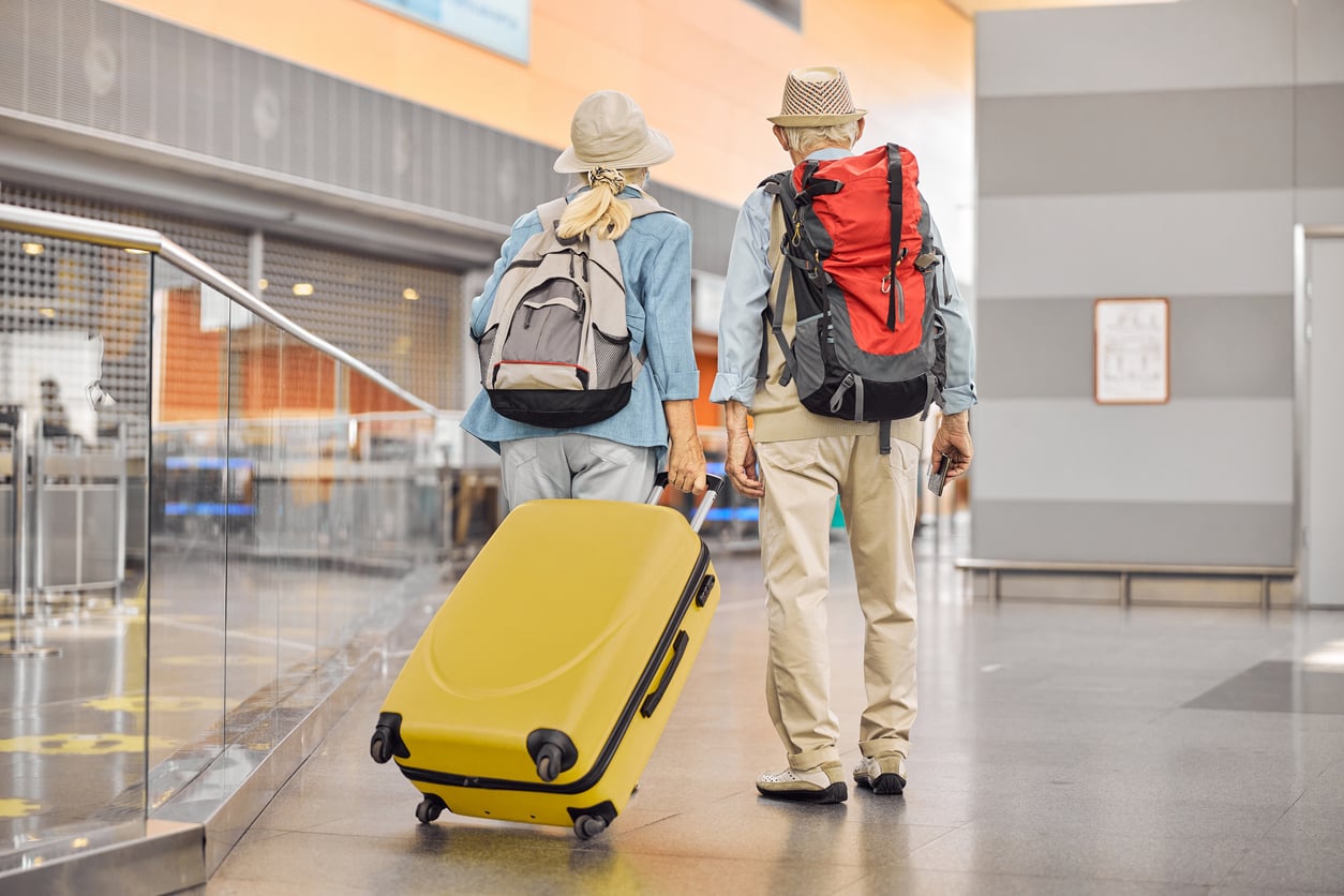 Two older adults with hats and backpacks walk side by side in an airport, one rolling a yellow suitcase. Reflecting distinct travel behaviors, they navigate the modern setting with ease, large windows and a sleek railing visible in the background.