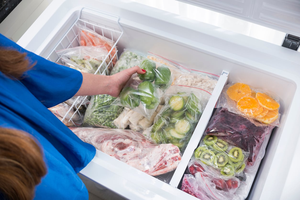 A person in a blue shirt places a bag of green bell peppers into a freezer filled with assorted frozen fruits, vegetables, and meat, all in clear plastic bags.