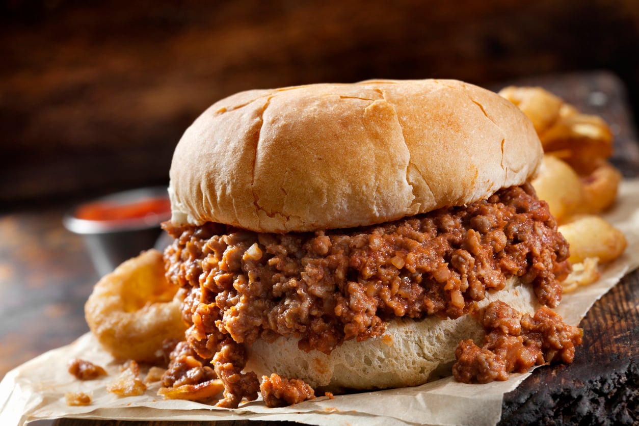 A sloppy joe sandwich with seasoned ground beef on a bun sits on parchment paper, evoking the comforting meals middle-class moms made, accompanied by golden onion rings and a cup of ketchup in the background.