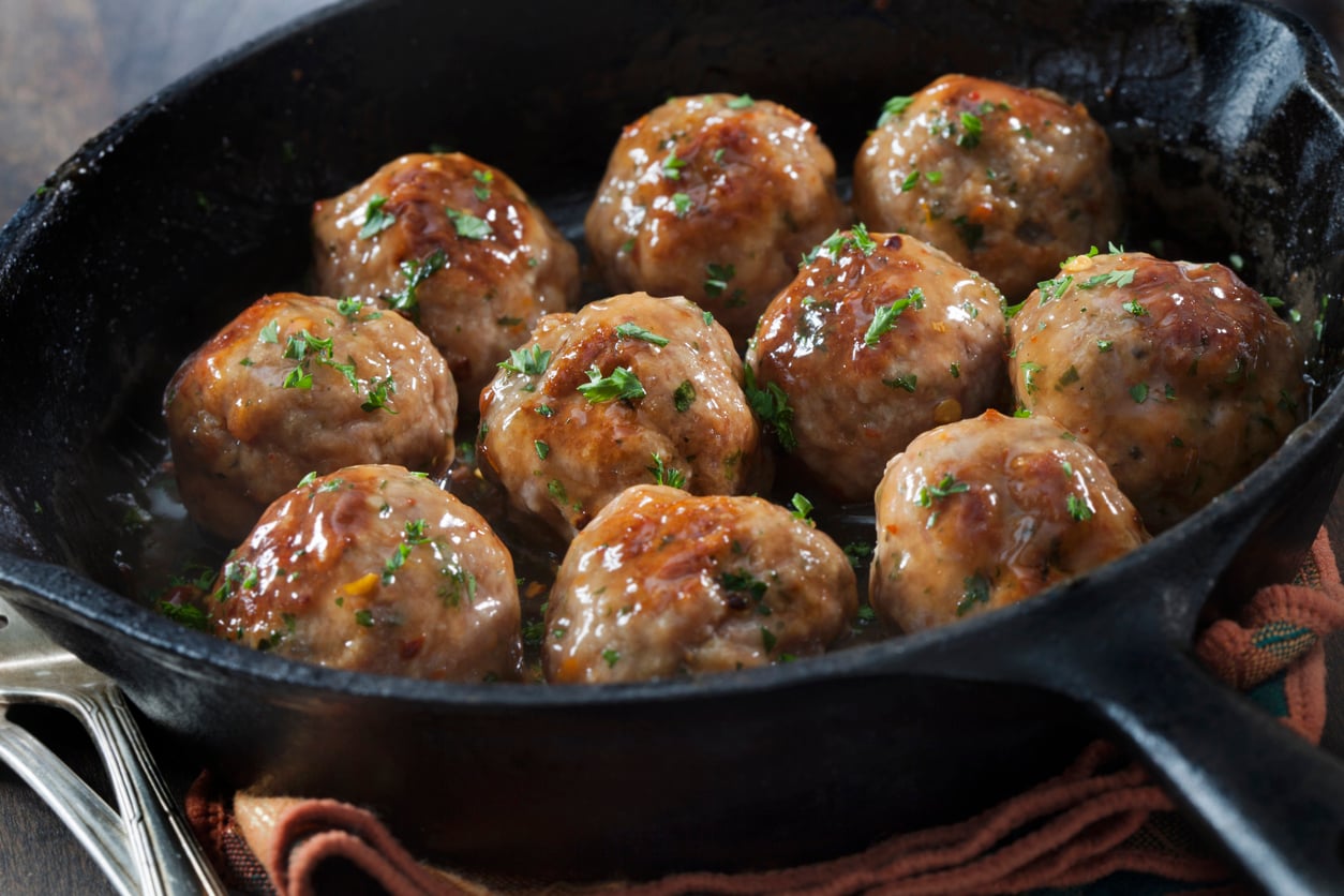 A cast iron skillet filled with glazed meatballs garnished with chopped herbs, resting on a cloth napkin.