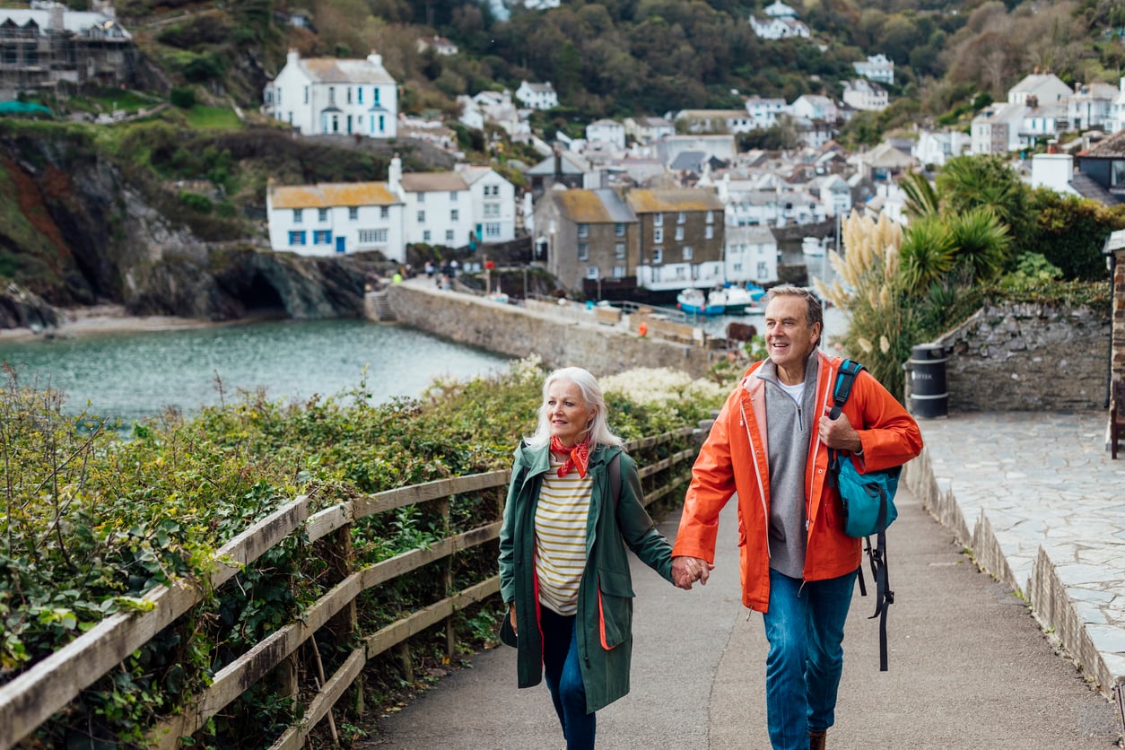 An older couple holding hands walks along a coastal path in a quaint village, smiling and dressed for cool weather—a scene that beautifully reflects their travel habits. White houses and green hills are visible in the background near the water.
