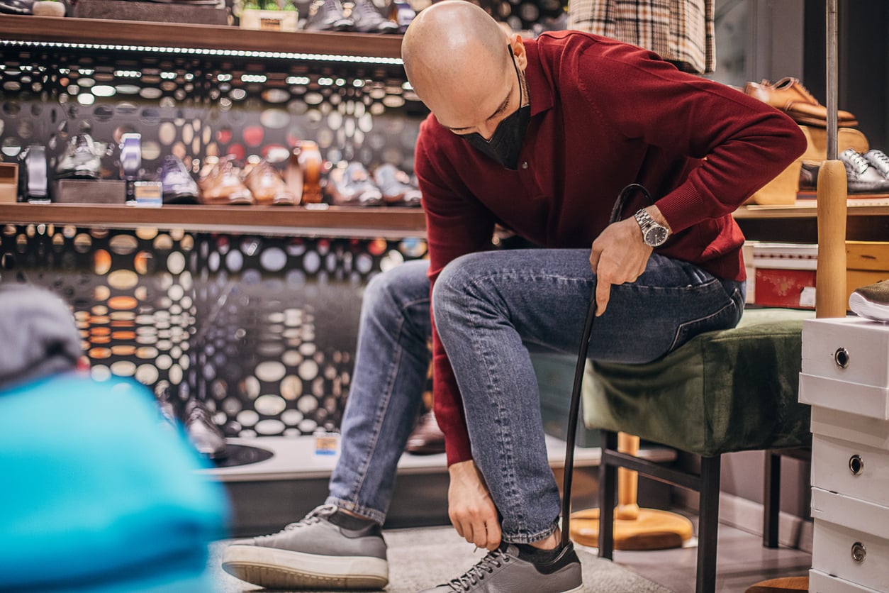 A man in a red sweater and jeans sits in a shoe store, using a shoehorn to put on gray sneakers. Shelves of shoes are visible behind him.