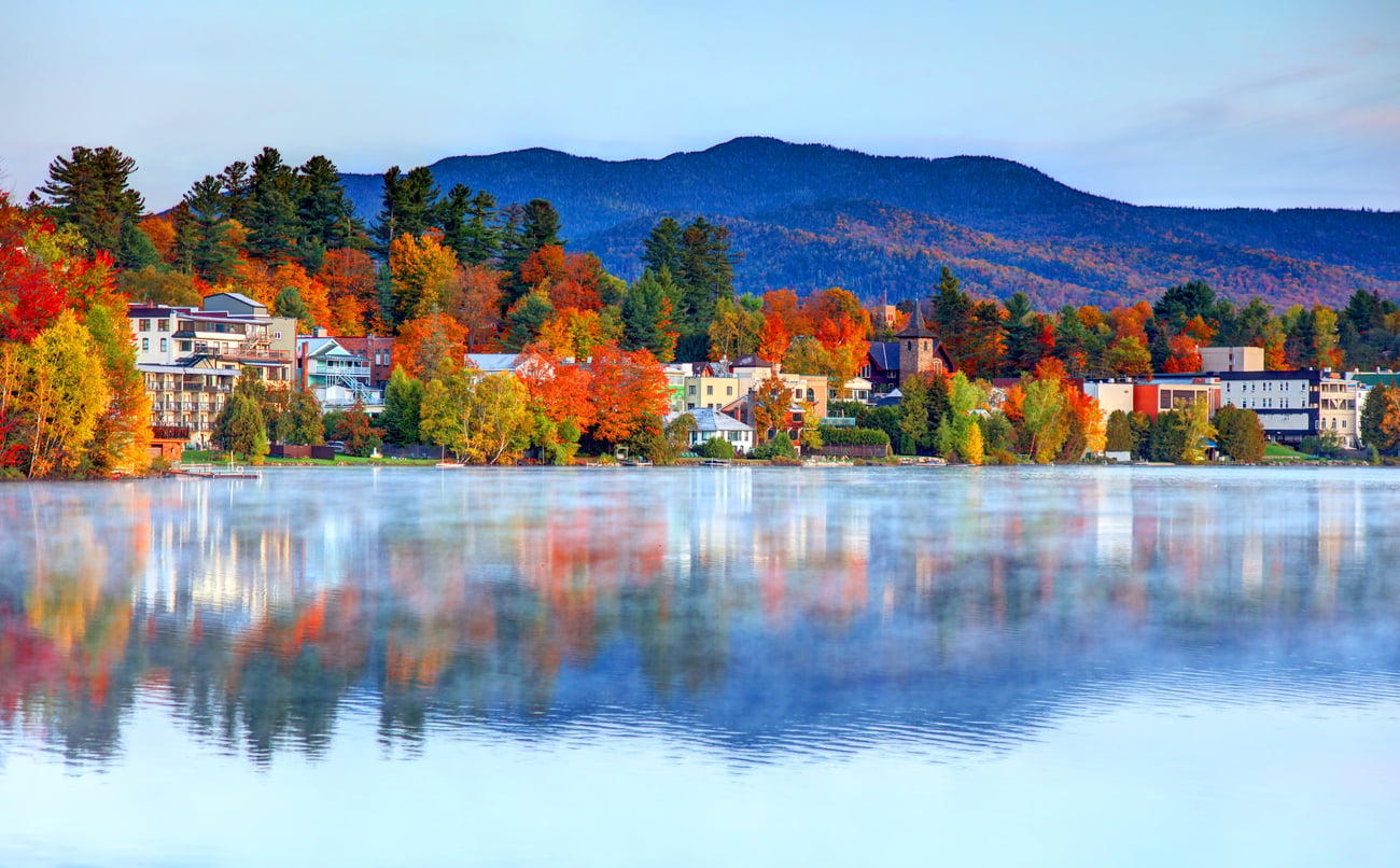 Colorful autumn trees and buildings line the shore of a calm lake, with reflections on the water and forested mountains in the background under a cloudy sky—capturing the charm often found in some of the best fall towns in America.