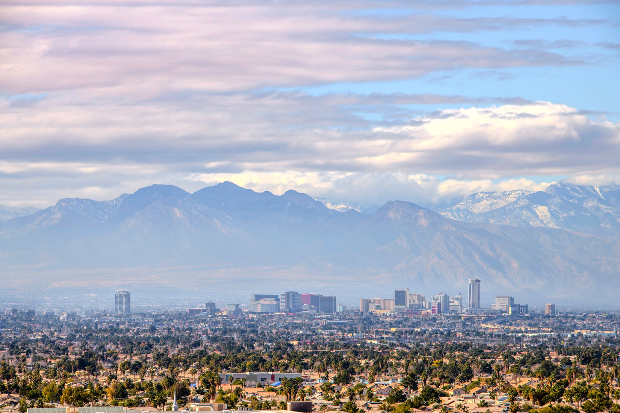 A city skyline with tall buildings is set against distant mountains under a partly cloudy sky, with a foreground of scattered houses and trees—much like the iconic vistas found in many Facts About Las Vegas, where urban landscapes meet stunning natural backdrops.