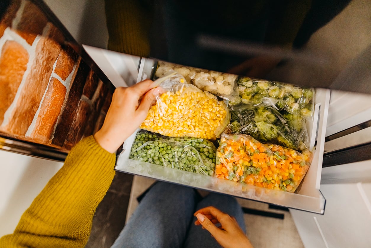 A person opens a freezer drawer filled with clear bags of frozen vegetables, including corn, peas, broccoli, and mixed vegetables. Only their hands and legs are visible.