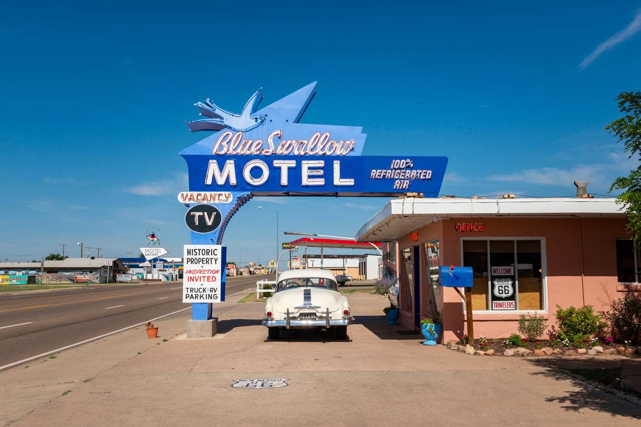 A vintage car is parked in front of the Blue Swallow Motel, one of the Classic Roadside Attractions on Route 66, with a large neon sign glowing under a clear blue sky. The motel has a retro look and a pink office building.