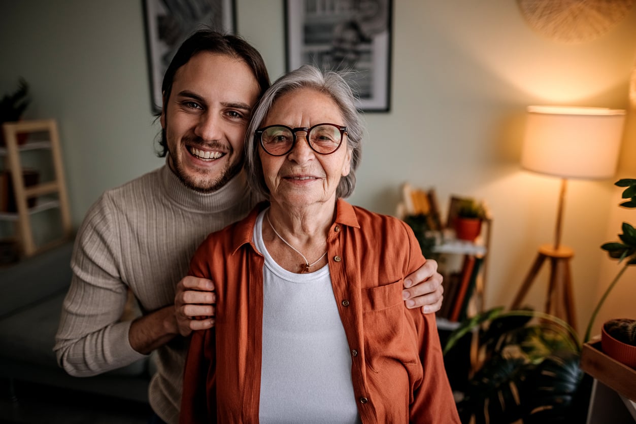 A younger man stands behind an older woman, both smiling at the camera in a cozy, well-lit living room with plants and warm decor—capturing the warmth and connection that reflect Baby Boomer changes across generations.