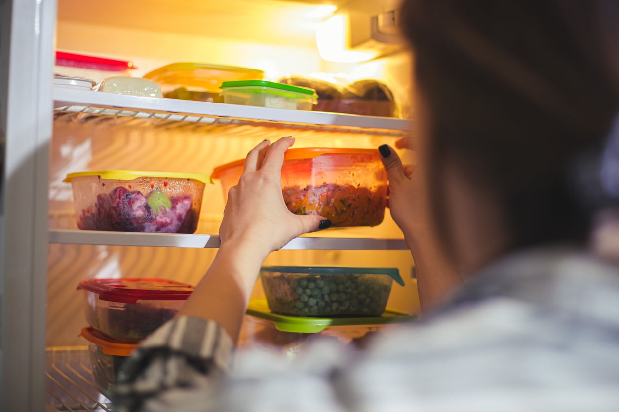 A person places a container of food inside a refrigerator filled with colorful, sealed containers holding various leftovers.