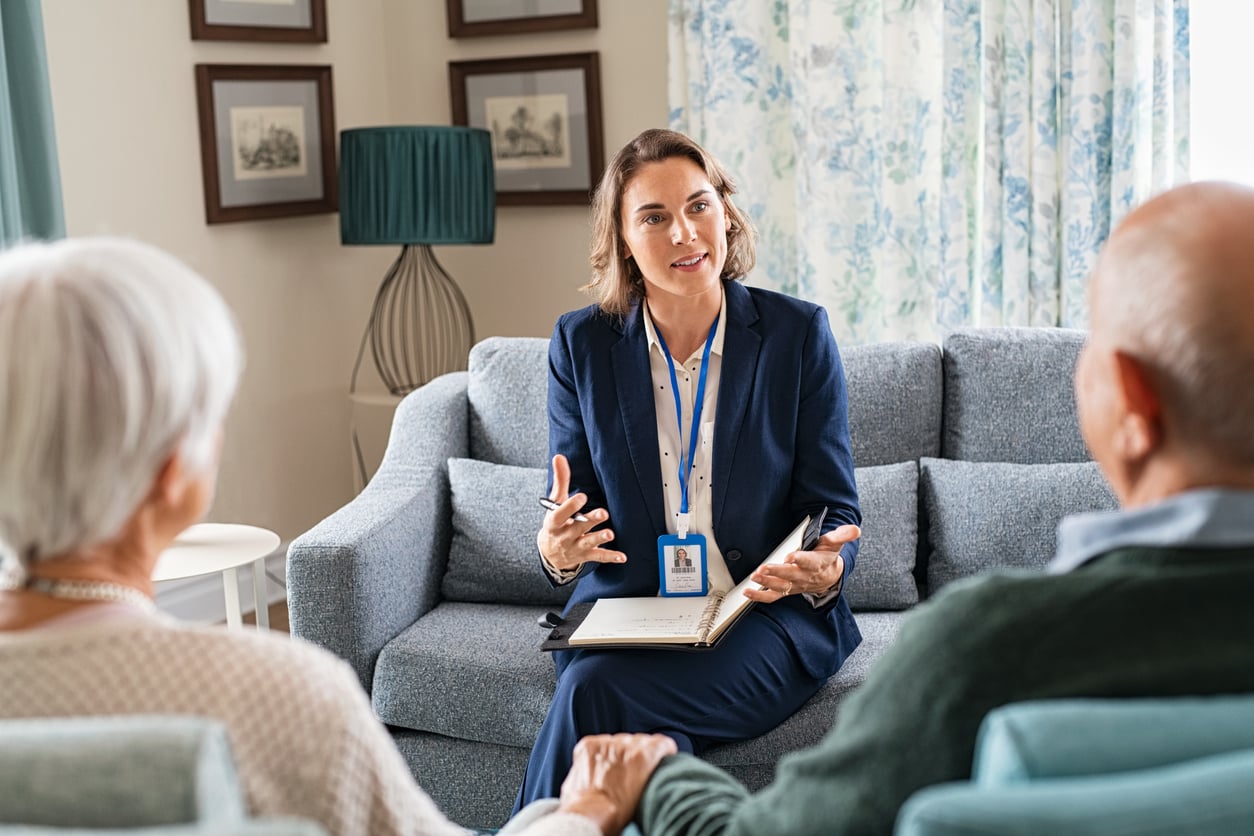 A woman in a suit sits on a couch, talking to an older couple who are holding hands. She has a notepad and wears a name badge, suggesting a professional meeting or counseling session in a cozy living room, where she may be offering retirement tips to help guide their future plans.