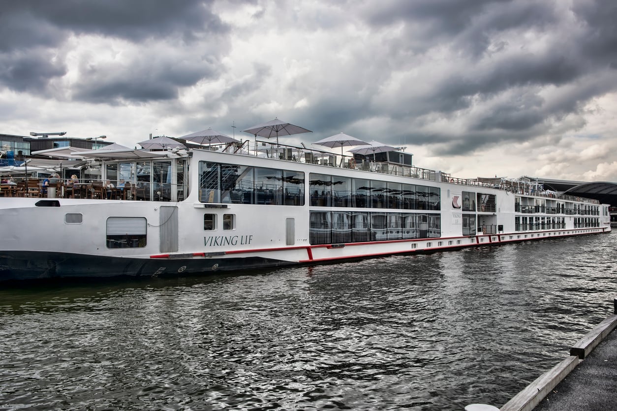A large white river cruise ship named “Viking Lif” is docked on the water under a cloudy sky, with outdoor seating and umbrellas visible on the upper deck.