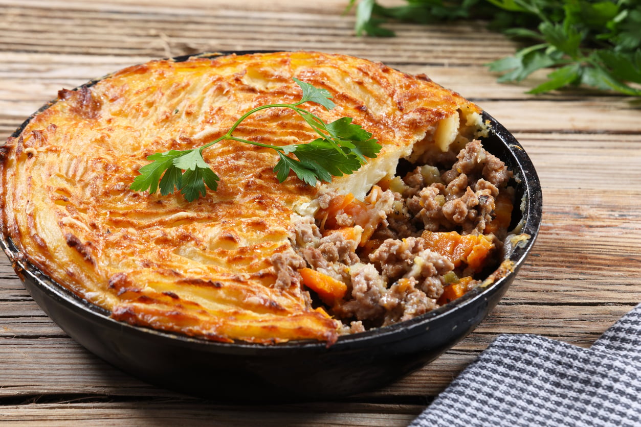 A baked shepherd’s pie in a black dish, topped with golden mashed potatoes, with a portion removed to reveal ground meat and vegetables inside. Parsley garnish on top and a checkered napkin nearby.