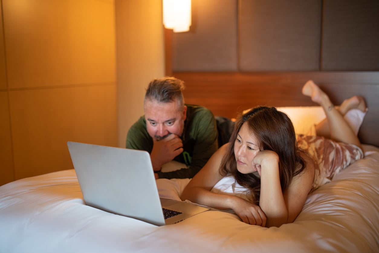 A man and woman lie on a bed in a cozy room, propped up on their elbows, watching something intently on a laptop placed in front of them.