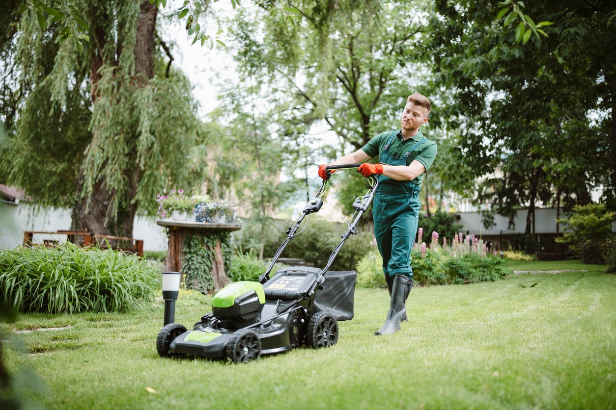 A person wearing green coveralls and boots mows a grassy lawn with a green and black lawnmower in a lush, landscaped garden surrounded by trees and plants.