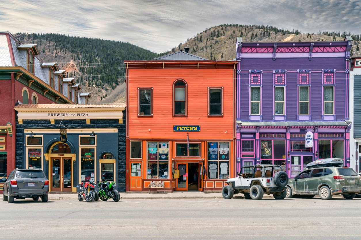 Colorful storefronts, including a brewery, line a small town street with cars and motorcycles parked in front—evoking the charm of nostalgic travel destinations. Hills covered in trees rise in the background under a cloudy sky.