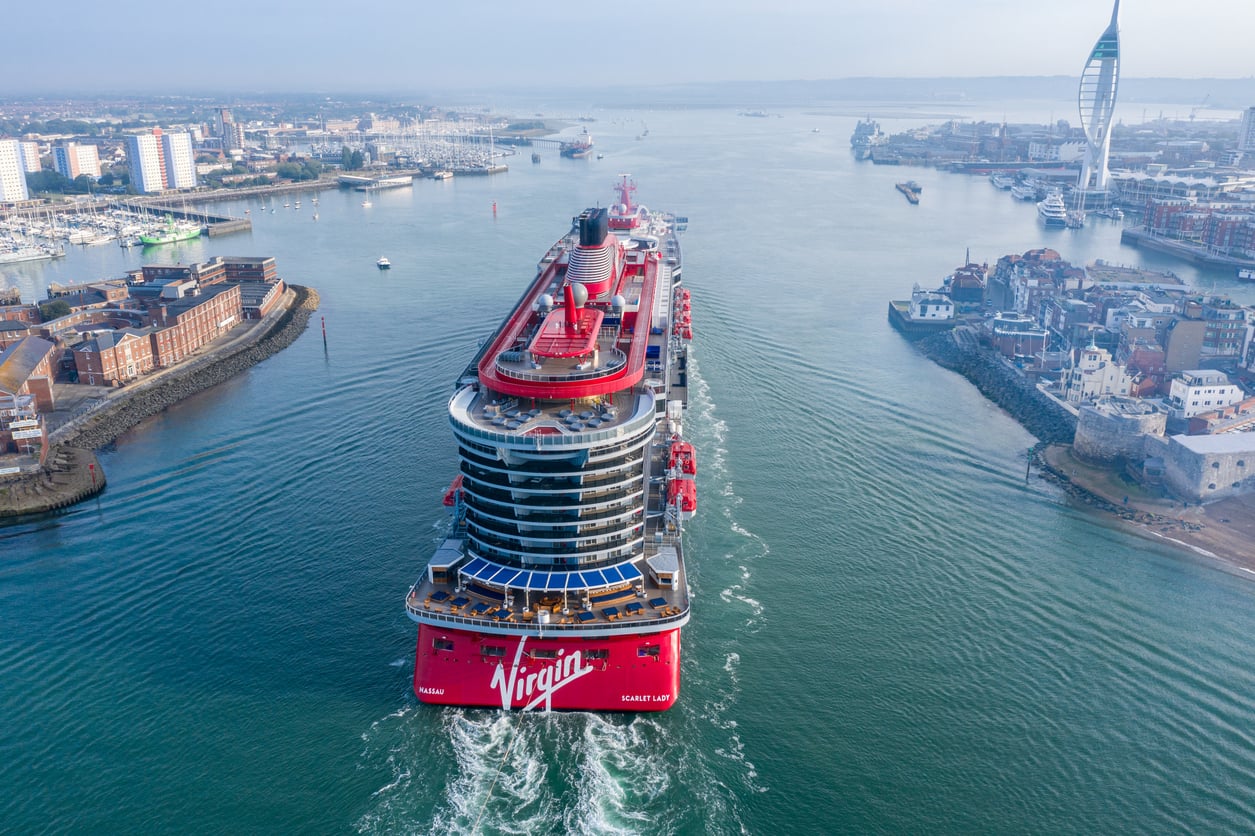 A large red cruise ship labeled “Virgin” sails from a harbor, with city buildings and a tall tower visible on both sides of the water under a clear sky.