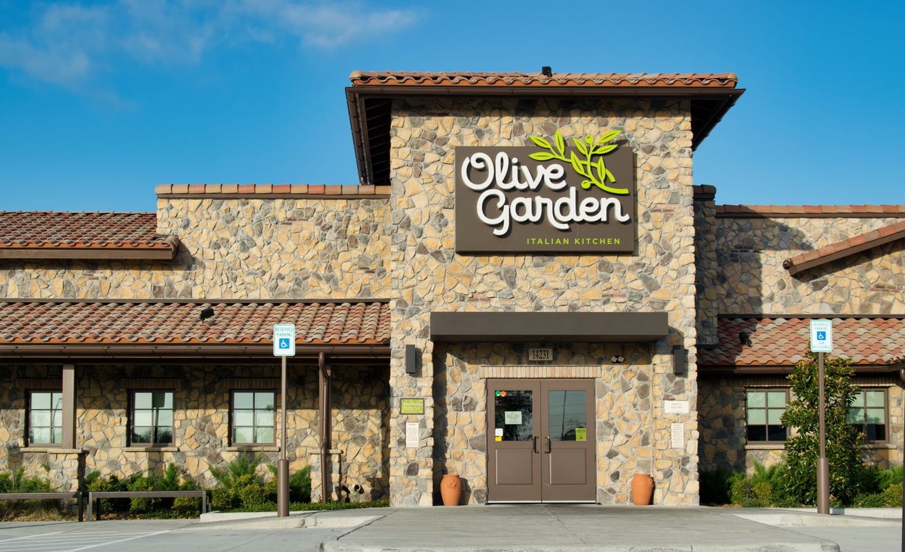 The exterior of an Olive Garden Italian Kitchen restaurant with a stone facade, large sign, and double doors, under a clear blue sky. Handicap parking spaces are visible in front.