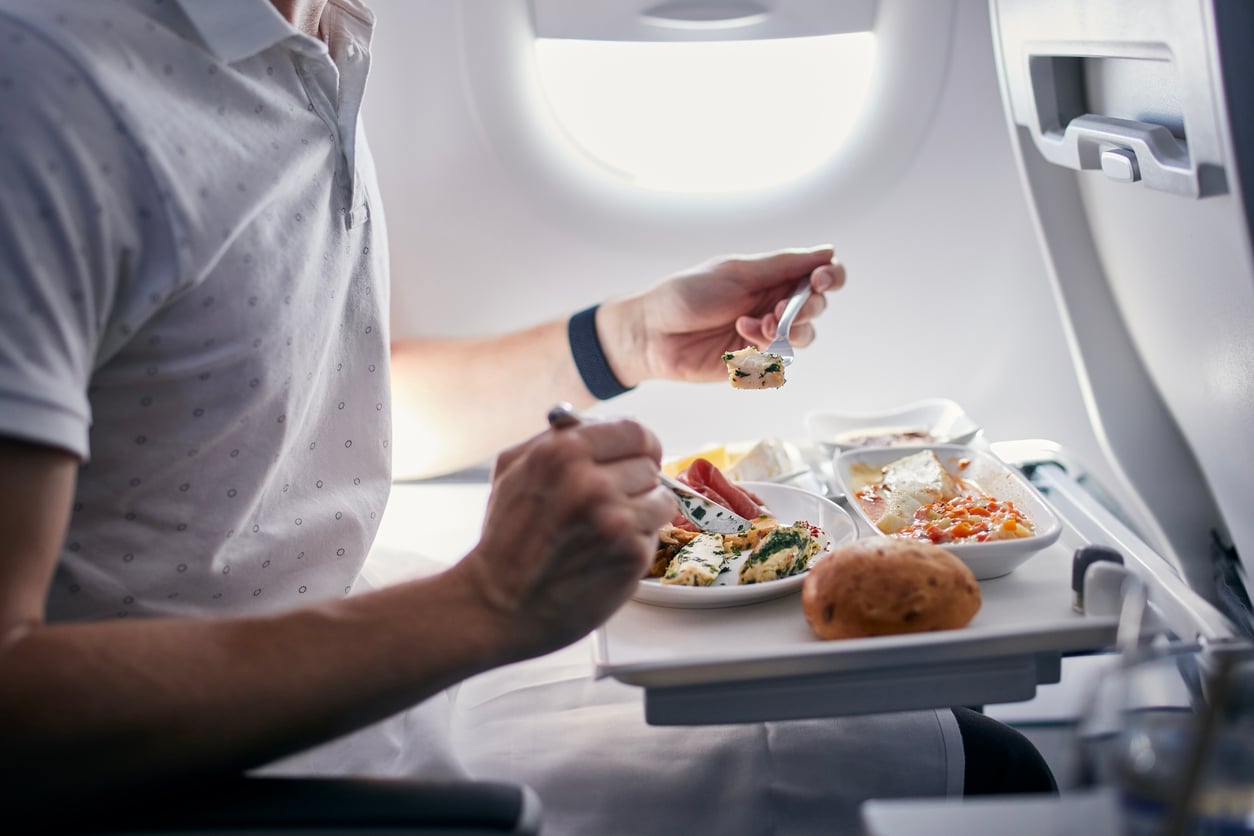 A person in a white shirt is eating a meal from a tray table on an airplane. The tray holds several dishes, including salad, bread, and a main course. Light shines through the airplane window.