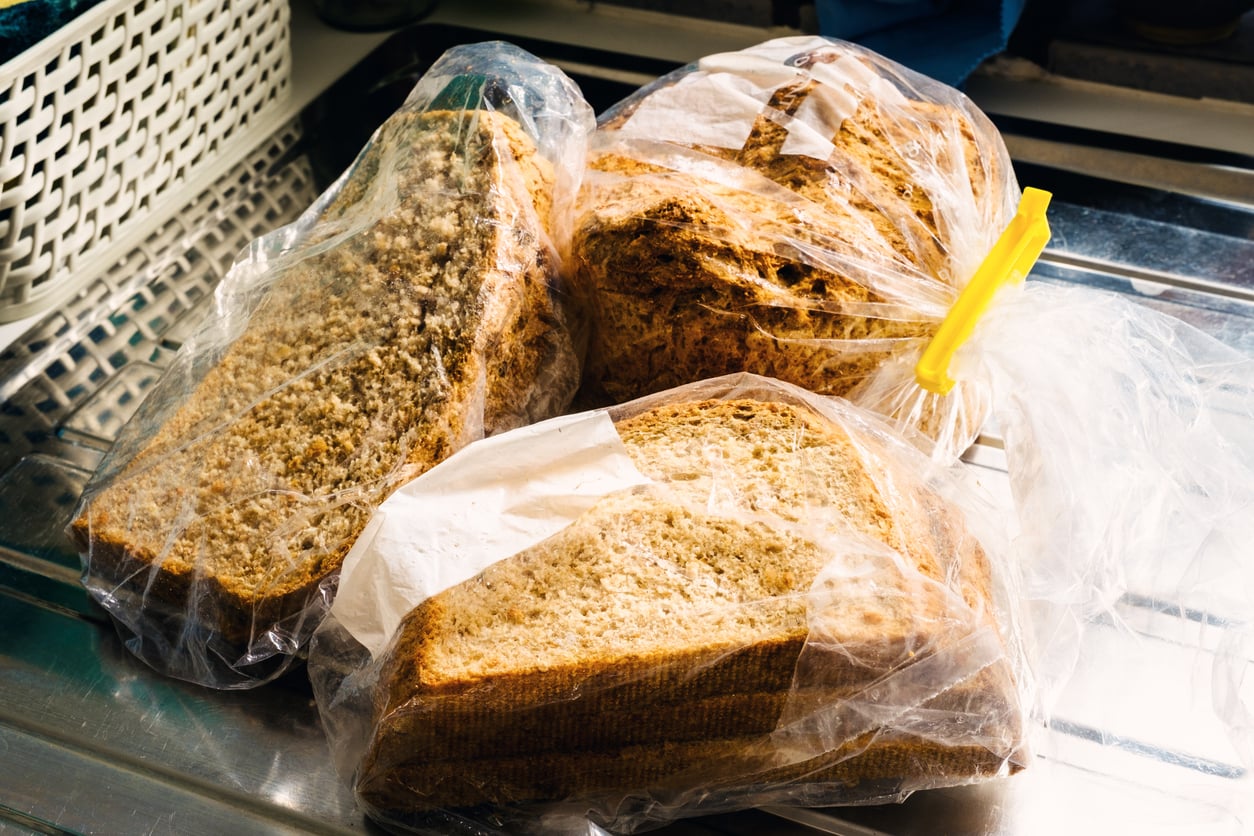 Three chunks of sliced whole grain bread are stored in a clear plastic bag, sealed with a yellow clip, and placed on a metallic surface near a white basket.