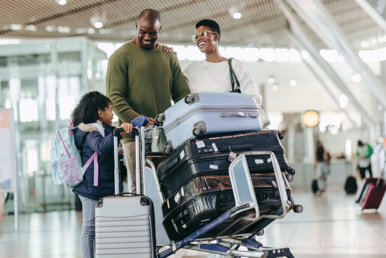 A smiling family with a young child stands in an airport, surrounded by suitcases and luggage on a cart, preparing to travel.