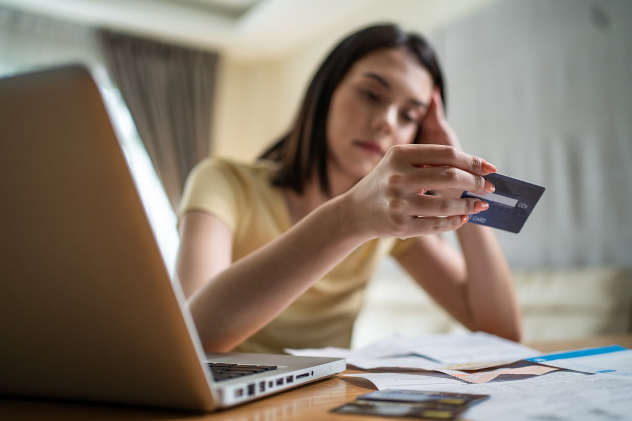 A woman sits at a table with a laptop, looking stressed while holding a credit card and reviewing bills or financial documents.