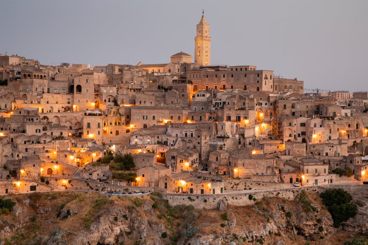 Historic stone buildings of Matera, Italy, are illuminated by warm lights at dusk. The structures are densely packed on a hillside with a church tower rising above the city against a soft evening sky.