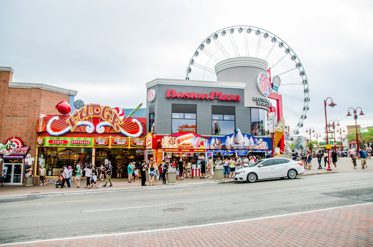 Colorful street scene with a Boston Pizza restaurant, Fudge shop, and large Ferris wheel in the background. People walk on the sidewalk, and a white car is parked on the road in front.