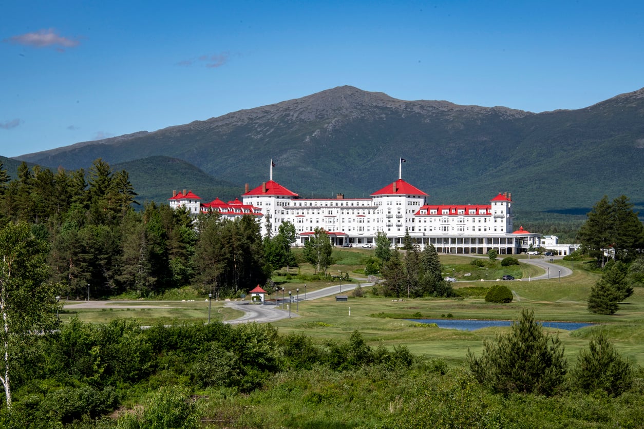 A large white hotel with red roofs sits among green trees and grass, with mountains rising in the background under a clear blue sky—a stunning example of Historic Hotels blending timeless elegance with breathtaking natural beauty.