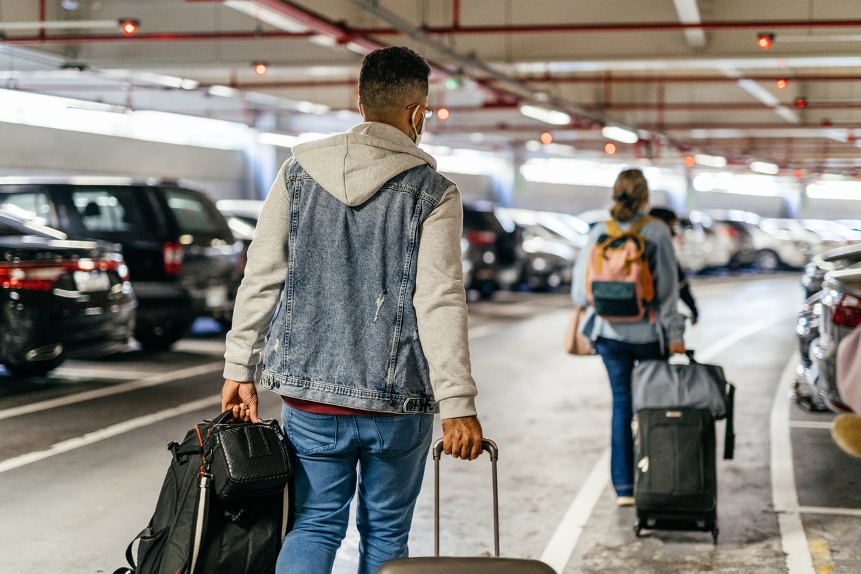 Two people walk through a parking garage, each pulling a suitcase and carrying bags, while surrounded by parked cars.