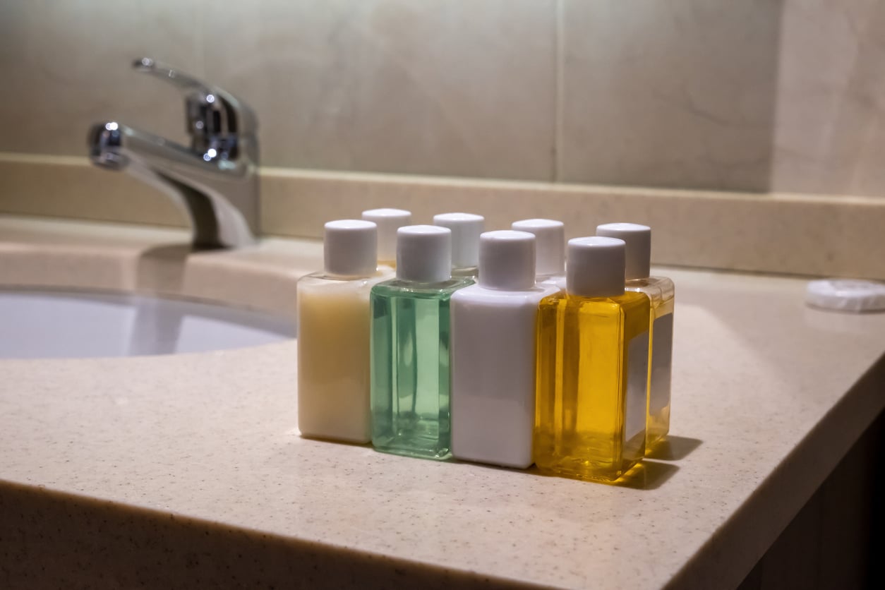 Six small bottles of toiletry liquids in various colors (white, green, yellow)—reminiscent of the things hotels used to offer guests—are lined up on a bathroom countertop near a sink and faucet.