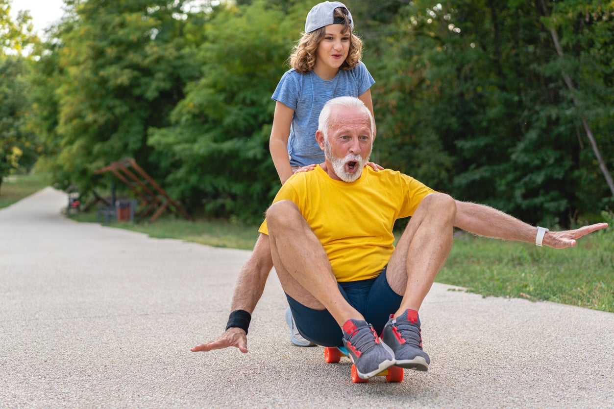 An older man in a yellow shirt rides a skateboard while a young girl, wearing a cap, stands behind him smiling. They are on a path surrounded by green trees, and both appear to be enjoying the fun moment.