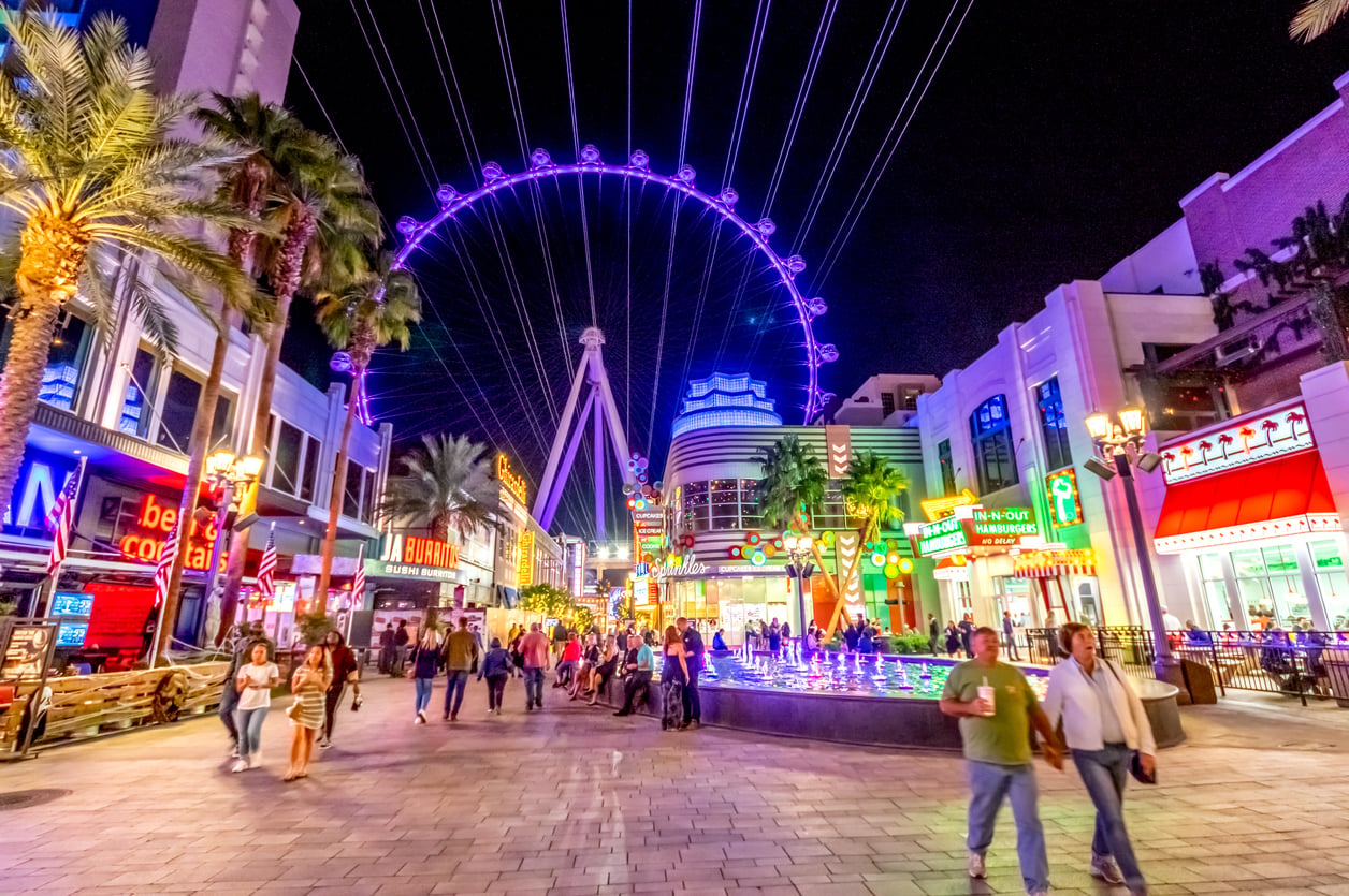 People walk along a lively, brightly lit outdoor plaza at night with neon signs and palm trees; a large illuminated Ferris wheel stands in the background against the night sky.