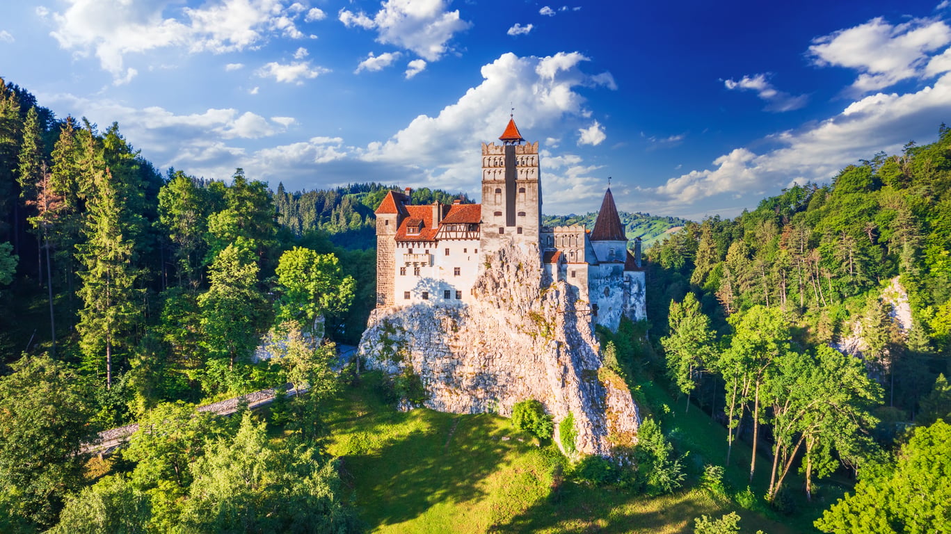 A large medieval castle with red roofs sits atop a rocky hill, surrounded by dense green forest under a bright blue sky with scattered clouds—one of those enchanting travel spots Boomers are learning to love.