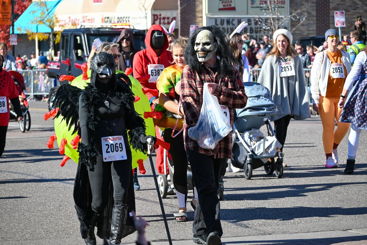 People in costumes participate in a parade or fun run, celebrating the festive spirit of Halloween. Visible costumes include a black-winged creature, a monster mask, a turtle, and others. Some push strollers, and many wear numbered race bibs. It’s a sunny day.