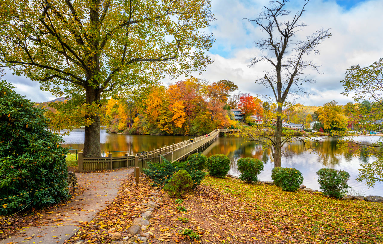 A path leads to a wooden bridge over a calm lake, surrounded by vibrant autumn trees with red, orange, and yellow leaves under a partly cloudy sky—capturing the picturesque charm often found in the best fall towns in America. Shrubs and fallen leaves line the walkway.