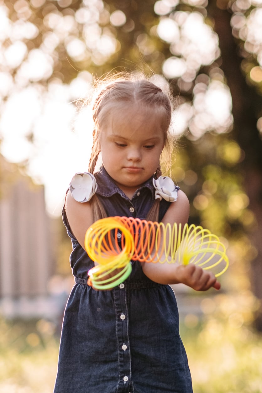 A young girl with braids and Down syndrome, wearing a denim dress, plays with a neon slinky outdoors in soft sunlight.
