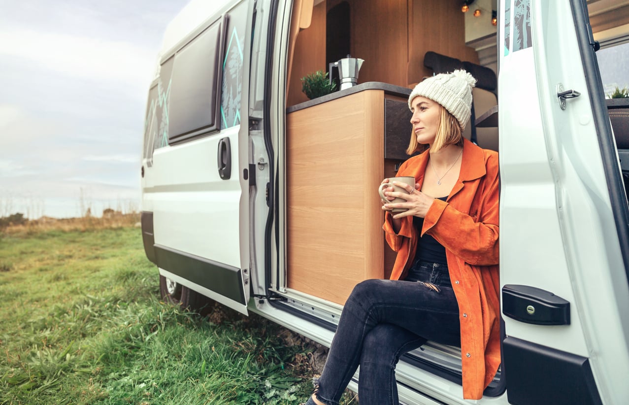 A woman in an orange coat and white beanie sits in the open doorway of a camper van, holding a mug and looking content. The van is parked on grass with a peaceful outdoor backdrop.