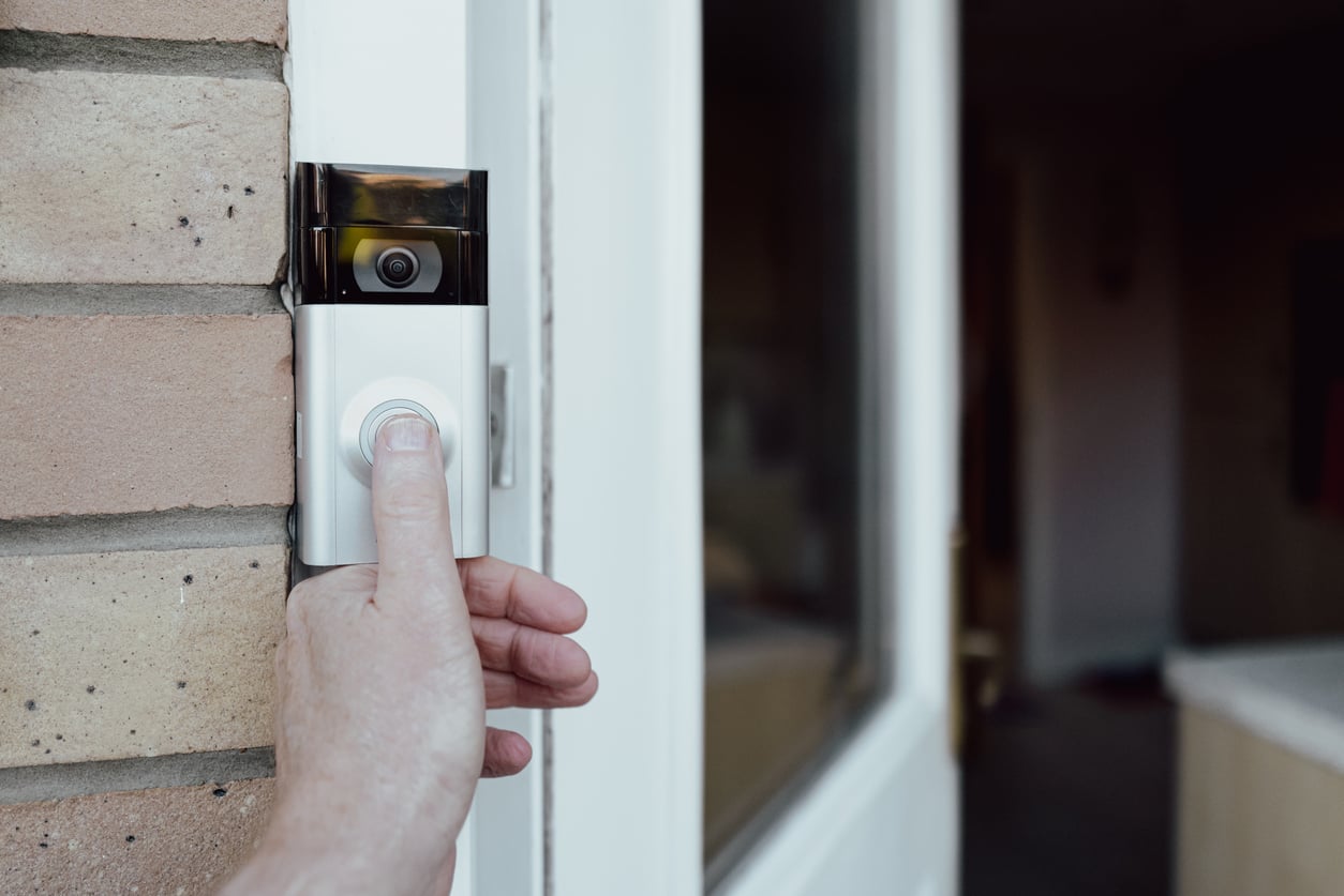 A person presses the button on a smart video doorbell mounted next to a front door on a brick wall.