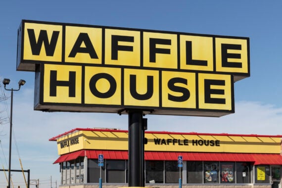 Large yellow Waffle House sign with black letters in front of a Waffle House restaurant building, featuring a red roof and blue sky in the background.